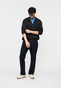 Homme portant des lunettes de soleil, un sweat-shirt noir à zip court avec le logo GAP, une chemise bleue, un jean foncé et des baskets blanches, debout les bras croisés.
