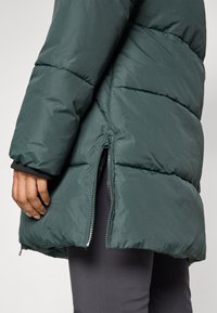 Abrigo acolchado en verde oscuro con un bolsillo con cremallera en el lateral y puños acanalados. Presenta una textura suave y un cuello alto para mayor calidez.