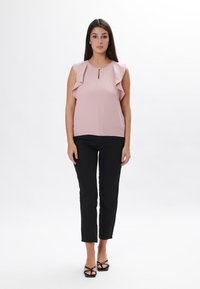 Blusa senza maniche di colore rosa chiaro con spalle arricciate e scollatura a chiave, abbinata a pantaloni neri aderenti e sandali neri con cinturino.