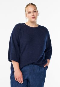 Suéter de punto azul marino con un ajuste holgado, mangas de ¾ de longitud y cuello redondo. Combinado con pantalones de mezclilla oscuros con cintura con cordón.