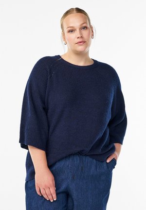 Navy blue knitted sweater with a loose fit, ¾ length sleeves, and a rounded neckline. Paired with dark denim pants featuring a drawstring waist.