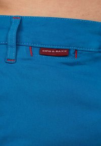 Blauwe katoenen broek met rode stiksels en een label met "CIPO & BAXX." Het ontwerp bevat een lus op de tailleband.