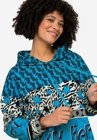 Kleurvolle capuchonjack met een mix van blauwe paisley-, dierenprint- en sterrenpatronen. De stof heeft een gladde textuur met contrasterende trekkoorden.