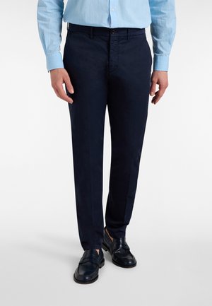 Uomo che indossa pantaloni eleganti blu scuro, camicia azzurro chiaro con bottoni e mocassini neri in pelle in piedi su sfondo bianco.