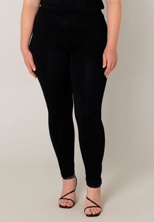 Vrouw die zwarte aansluitende leggings en zwarte hoge hakken met bandjes draagt, staand tegen een effen lichte achtergrond.