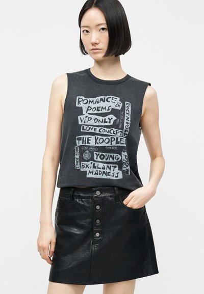 Νεαρή γυναίκα με κοντά μαύρα μαλλιά, ντυμένη με αμάνικο μαύρο μπλουζάκι με σχέδιο και μαύρη δερμάτινη φούστα με κουμπιά μπροστά, ποζάρει με το ένα χέρι στην τσέπη.