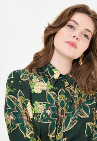 Green floral blouse with a collar, featuring yellow and pink roses, intricate leaf patterns, and buttons. Fabric has a smooth texture.