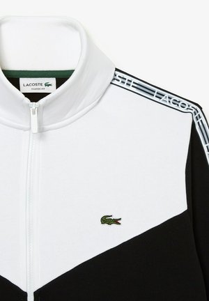 Veste zippée noire et blanche avec un col montant. Présente une texture côtelée, un logo brodé vert sur la poitrine, et des accents rayés sur les épaules.