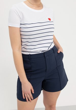 Kobieta w białym t-shircie z granatowymi poziomymi paskami i małym czerwonym sercem, do tego dopasowane granatowe szorty z wysokim stanem.