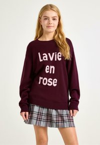 Bordeaux stickad tröja med texten "Lavie en rose" i ljusrosa, med en ribbad rund hals och långa ärmar, i kombination med en rutig kjol.