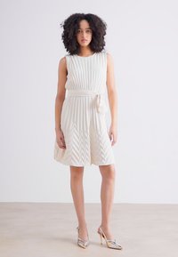 Pleated cream dress with sleeveless design, cinched waist and accentuating bow. Features chevron pattern and flared skirt. Paired with silver heels.