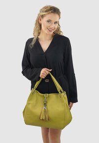 Groene leren handtas met twee handvaten, een kwastdetail, zilveren hardware, een zachte textuur en een ronde vorm. G gedragen met een zwart outfit.