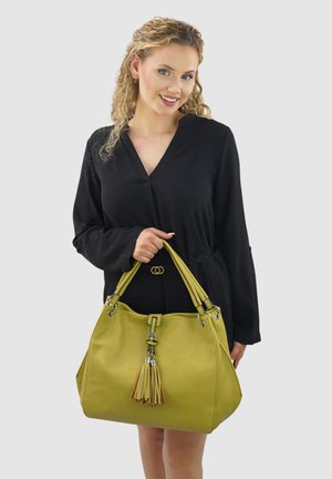 Grüne Lederhandtasche mit zwei Griffen, Quaste, silbernen Details, weicher Textur und runder Form. Getragen mit einem schwarzen Outfit.