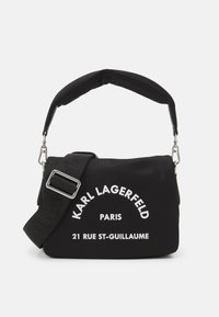 Czarna torba na ramię wykonana z miękkiego materiału, z wyściełanym paskiem, srebrnymi detalami oraz białym nadrukiem z nazwą marki i adresem.