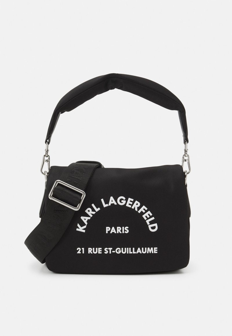 Czarna torba na ramię wykonana z miękkiego materiału, z wyściełanym paskiem, srebrnymi detalami oraz białym nadrukiem z nazwą marki i adresem.