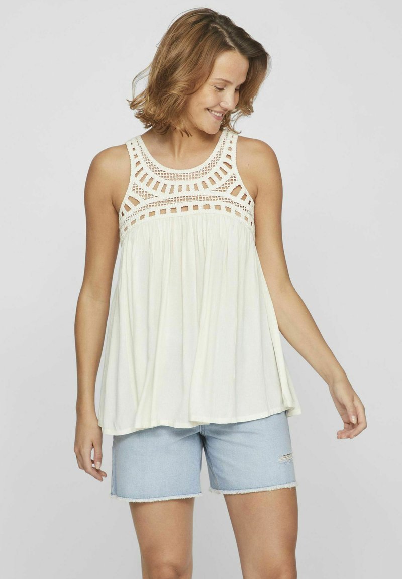 Top sin mangas de color crema claro con intrincados detalles de croché en el escote. Tejido suave y fluido con un ajuste holgado. Combinado con pantalones cortos de mezclilla claros.