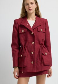 Giacca trapuntata rossa con design doppiopetto, bottoni dorati, due tasche frontali e vita con coulisse. Abbinata a una gonna con texture.