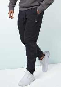 Schwarze Jogginghose mit schlankem Schnitt, ausgestattet mit seitlichen Reißverschlusstaschen und einem strukturierten Stoff. Getragen mit hellgrauen Sportschuhen.