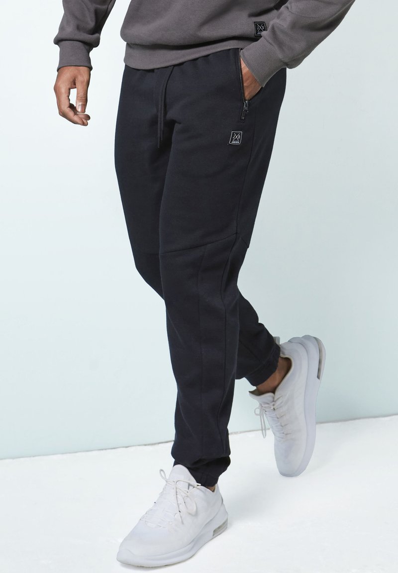 Schwarze Jogginghose mit schlankem Schnitt, ausgestattet mit seitlichen Reißverschlusstaschen und einem strukturierten Stoff. Getragen mit hellgrauen Sportschuhen.
