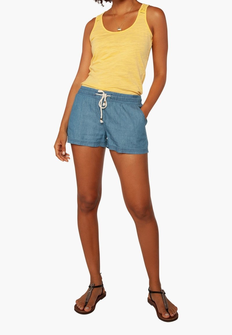 Keltainen hihaton paita, jossa on teksturoitua kangasta, yhdistettynä vaaleansinsisiin farkkushortseihin, joissa on narukuja. Mustat litteät sandaalit täydentävät asua.