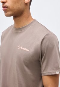 Άτομο που φοράει μπλουζάκι Berghaus σε αποχρώσεις του ταούπ με λογότυπο στο στήθος και το μανίκι, εμφανιζόμενο από τον ώμο μέχρι το πηγούνι σε απλό φόντο.