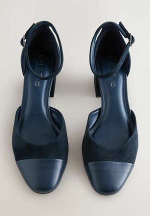 Scarpe col tacco nere in camoscio con una tomaia in camoscio e una punta in pelle liscia; presentano un cinturino alla caviglia con una fibbia argentata.