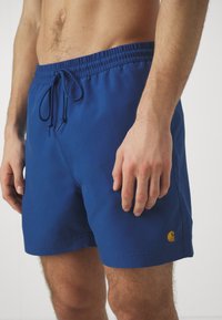 Bañador azul hecho de una tela suave, con una cintura elástica con cordón y un detalle de logotipo dorado en la parte inferior izquierda.