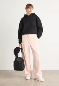 Sweatshirt à capuche noire, pantalon de survêtement rose, baskets blanches, casquette noire à la main, sac noir avec un détail en matériel circulaire, design et coupe décontractés.