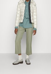 Person iført en beige pufferjakke med teksten "ANIMAL FREE", grønstribet skjorte, salvie cropped bukser, hvide sokker og grå loafers.