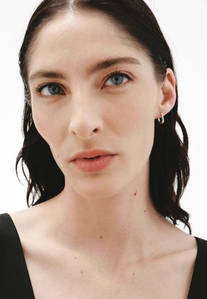 Primer plano de la cara de una persona con cabello oscuro y lacio, piel clara, ojos azules, pecas sutiles y pequeños pendientes de aro de plata.