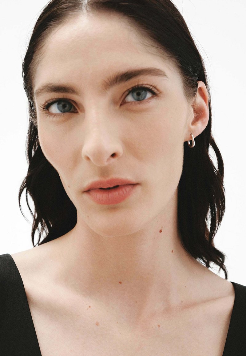 Primer plano de la cara de una persona con cabello oscuro y lacio, piel clara, ojos azules, pecas sutiles y pequeños pendientes de aro de plata.