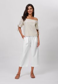 Off-Shoulder-Bluse in hellem Beige mit dezenten Streifen; kombiniert mit weißen, weit geschnittenen Hosen; Model trägt braune Sandalen.