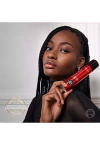 Femme aux cheveux tressés tenant près du visage le spray rouge L'Oréal Infallible 24H Setting Mist, portant un haut noir transparent, expression neutre.