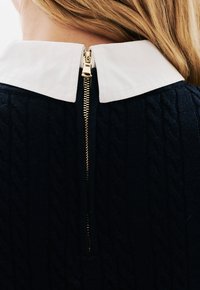 Marineblauer Kabelstrickpullover mit weißem Kragen, ausgestattet mit einem goldenen Reißverschluss auf der Rückseite. Die Textur ist gerippt und kunstvoll gemustert.