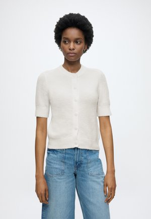 Femme aux cheveux courts et bouclés portant un cardigan blanc à manches courtes boutonné et un jean taille haute bleu clair, debout devant un fond uni.
