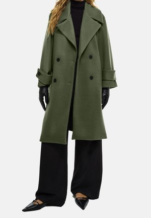 Kobieta ubrana w oversize oliwkowo-zielony płaszcz z podwójnym rzędem guzików, czarne szerokie spodnie, czarny golf, czarne rękawiczki skórzane i czarne szpiczaste baleriny.