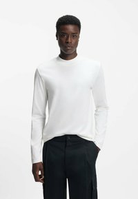 Langärmliges weißes T-Shirt aus leichtem Stoff, mit Rundhalsausschnitt und schmaler Passform, kombiniert mit schwarzen Hosen.