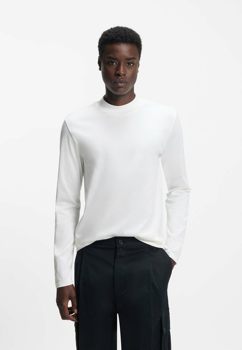 Langärmliges weißes T-Shirt aus leichtem Stoff, mit Rundhalsausschnitt und schmaler Passform, kombiniert mit schwarzen Hosen.