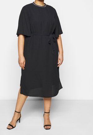 Frau trägt ein schwarzes Midikleid mit kurzen Ärmeln, einem Taillengürtel und schwarzen, hochhackigen Sandalen mit Knöchelriemen, steht vor einem weißen Hintergrund.