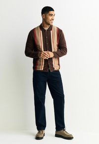 Barna csíkos kardigán piros, bézs és krém függőleges vonalakkal; gombos elülső kialakítás. Tengerészkék nadrággal és világos színű cipővel párosítva.