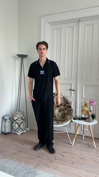 Jeune homme en tenue noire debout dans une pièce aux murs blancs, sol en bois, lanternes décoratives, une chaise recouverte de fourrure et une petite table avec des sculptures.