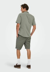 Chemise olive à manches courtes avec col, associée à un short gris hauteur genoux, à la fois structuré et décontracté. Chaussures brunes, chaussettes blanches.