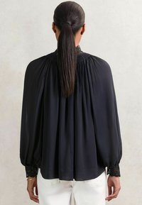 Schwarze Bluse aus leichtem Material mit gerafftem Ausschnitt, puffärmeligen langen Ärmeln und mit Spitze verzierten Bündchen, fließendes Design.