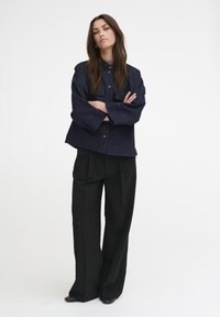 Giacca di jeans blu navy con chiusura a bottone e due tasche frontali, abbinata a pantaloni neri a gamba larga e scarpe nere. Tessuto dalla consistenza liscia.