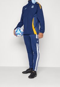 Marine trainingspak met gele accenten, lichte stof, jack met rits aan de voorkant, witte gestreepte broek, zwarte voetbalschoenen, en vastgehouden een meerkleurige voetbal.