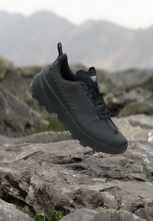 Schwarzer Dynafit Trail-Laufschuh mit robuster Sohle schwebt über felsigem Gelände mit verschwommenem Berg-Hintergrund.