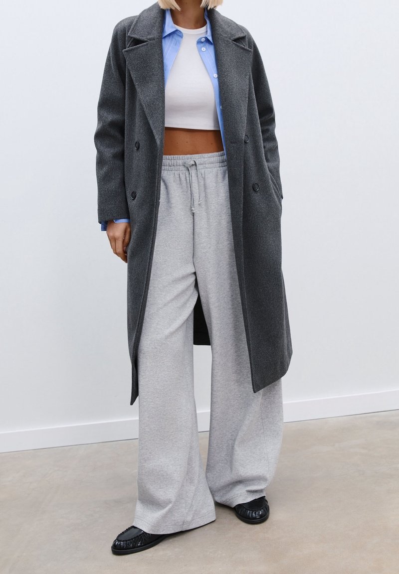 Charcoal gray double-breasted coat layered over a light blue shirt and white crop top, paired with light gray wide-leg pants and black flats.