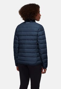 Marineblauwe pufferjack met een gequilt patroon, hoge kraag en logo op de achterkant. Het materiaal lijkt glad en geïsoleerd voor warmte.