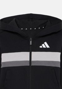 Crna zip-up hoodica s bijelim i sivim horizontalnim prugama te Adidas logotipom na lijevoj strani prsa i podstavljenim kapuljačom.