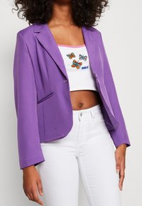 Purpurový blejzr s upraveným střihem, zapínání na jeden knoflík a dvěma předními kapsami. Nositý přes bílý crop top s barevnou výšivkou motýlů.